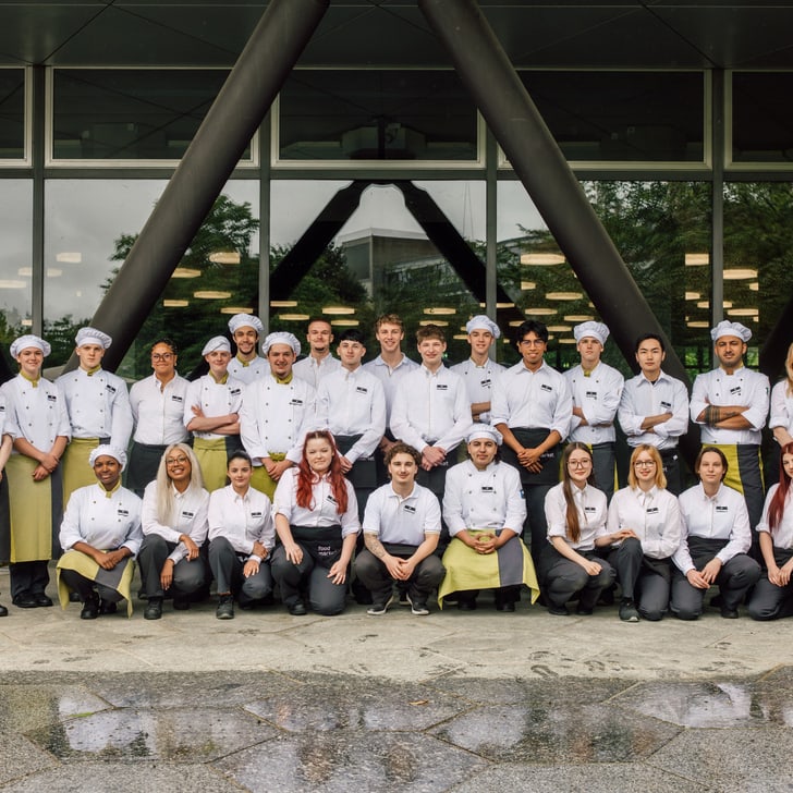 Gruppe von Köchen und Mitarbeitern vor einem Gebäude mit Glasfassade.