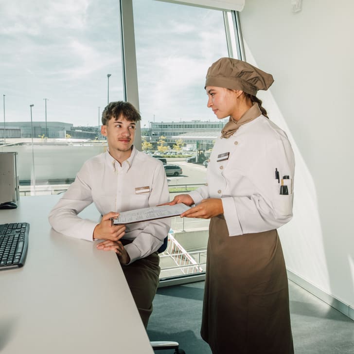 Zwei Personen in Arbeitsuniformen, eine sitzt am Schreibtisch, eine steht mit Tablet in Bürofensternähe.
