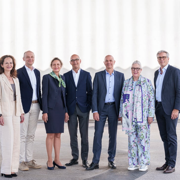 Gruppe von sieben Menschen in formeller Kleidung vor einer weissen Wand.