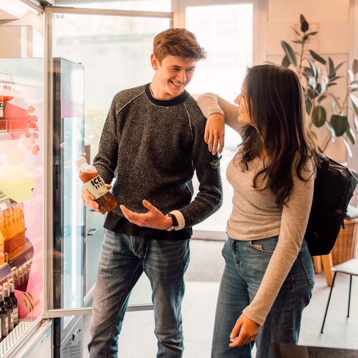 Zwei Personen stehen lächelnd vor einem offenen Kühlregal mit Getränken.