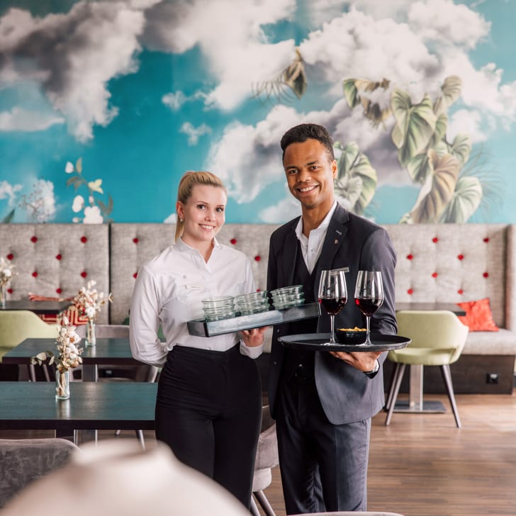 Zwei Kellner mit Tablett und Weingläsern in einem modernen Restaurant mit Wandmalerei.",