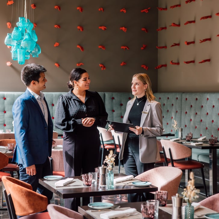 Drei Personen in einem Restaurantgespräch nahe gedeckter Tische.