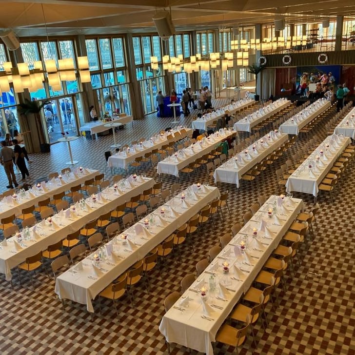 Grosser Saal mit langen gedeckten Tischen und Personen im Hintergrund.