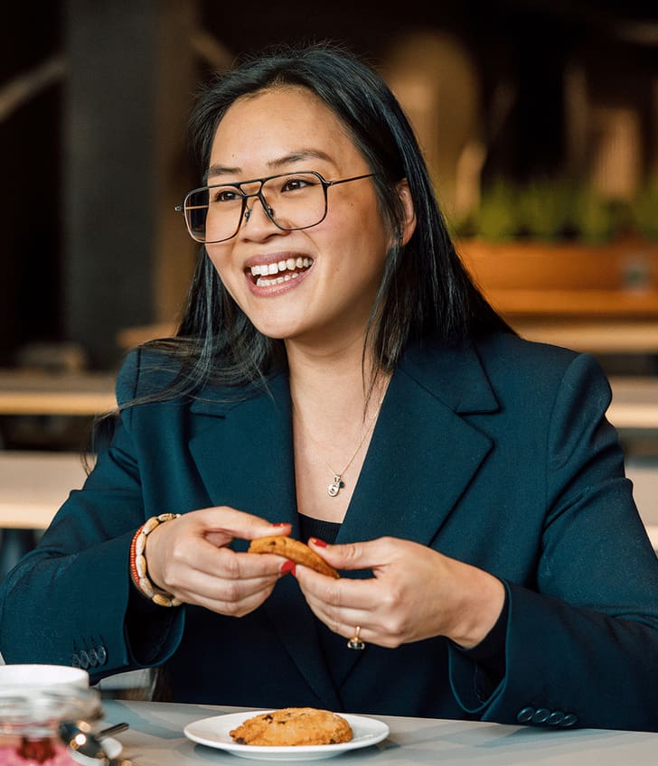Frau in Blazer lächelnd und Keks haltend am Tisch sitzend.