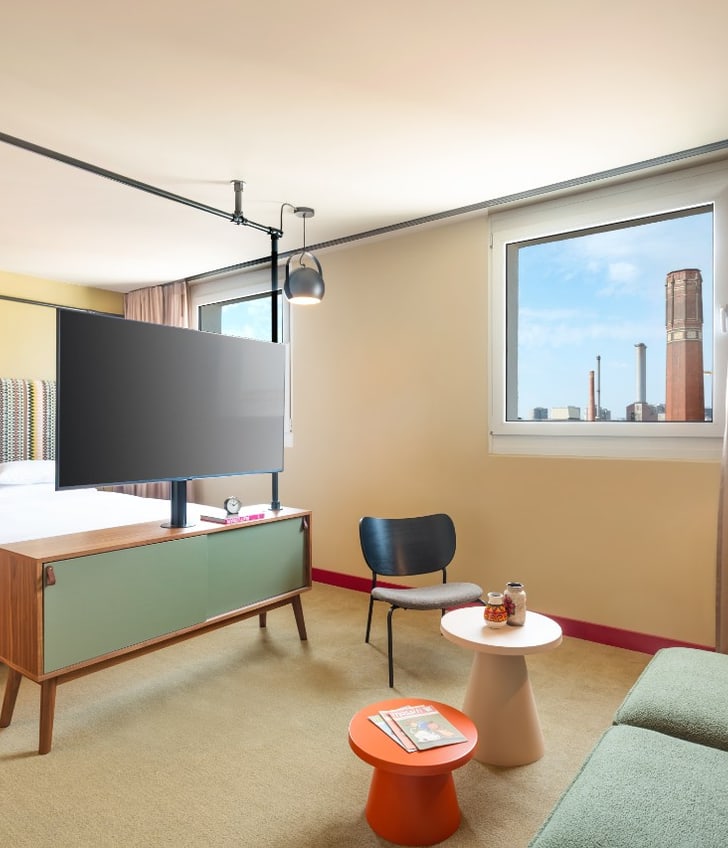 Hotelzimmer mit Doppelbett, Sofa, TV und Fensterblick auf Stadtgebäude.