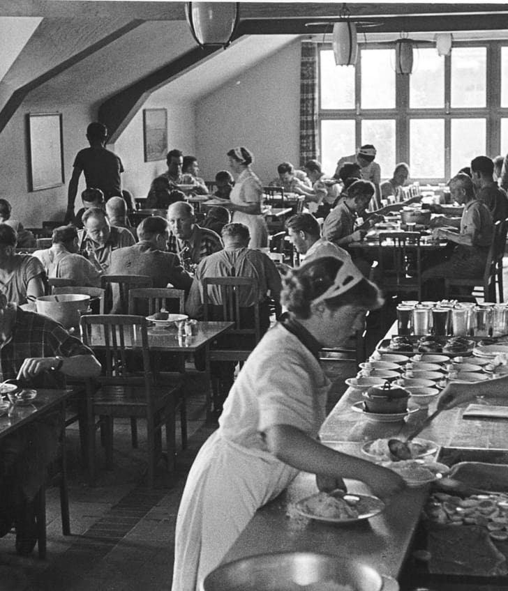 Historisches Foto eines Personalrestaurants in schwarz-weiss, wo viele Menschen beim Mittagessen sitzen und im Vordergrund an der Theke geschöpft wird.