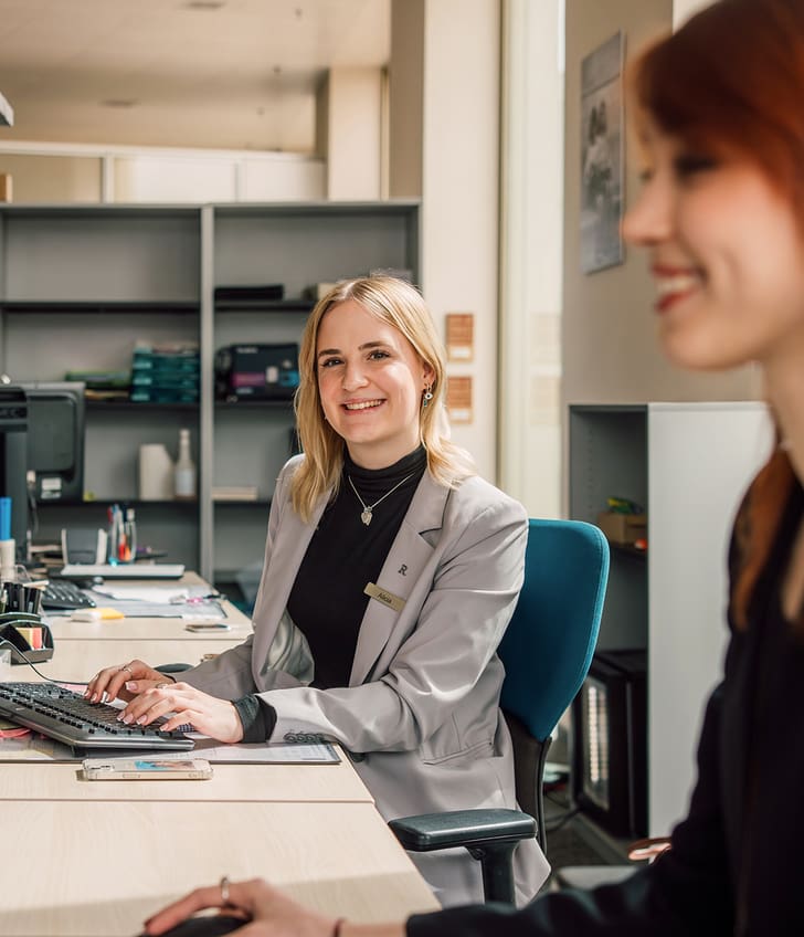 Zwei Frauen an Büroarbeitsplätzen, eine lächelnd zur Kamera.