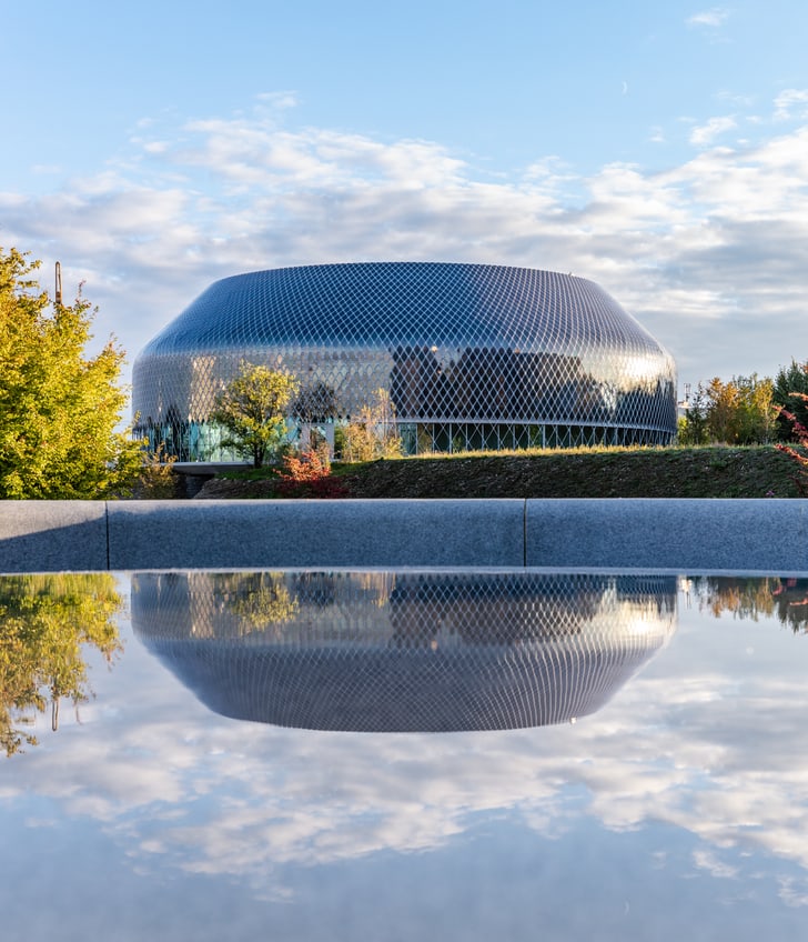 Modernes Gebäude mit Wasserreflexion und Bäumen im Vordergrund.