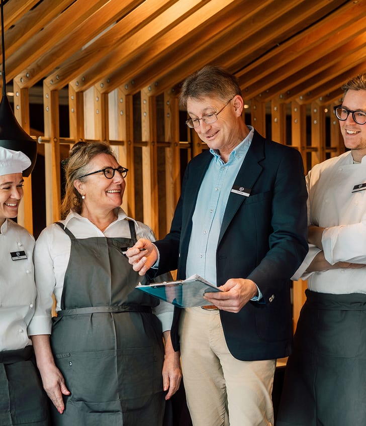 Drei Köche und ein Mann mit Klemmbrett im Gespräch in einer Küche.