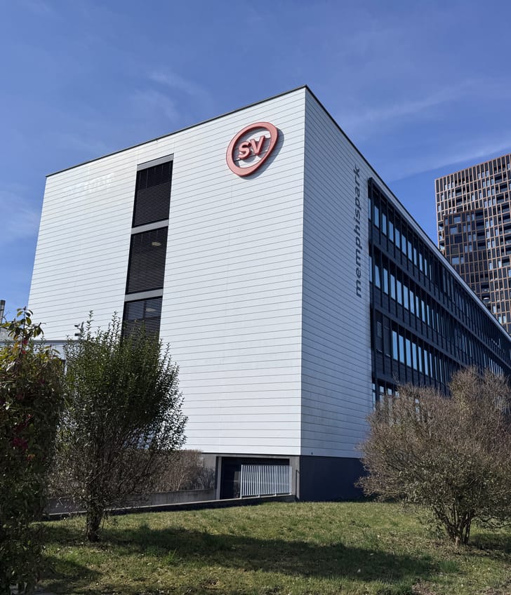 Bürogebäude mit SV-Logo, blauem Himmel, grüner Umgebung.
