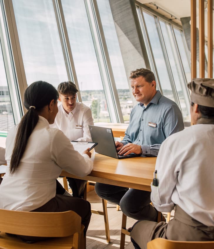 Leute in einem Meeting am Tisch mit Laptop und Fenstern im Hintergrund.