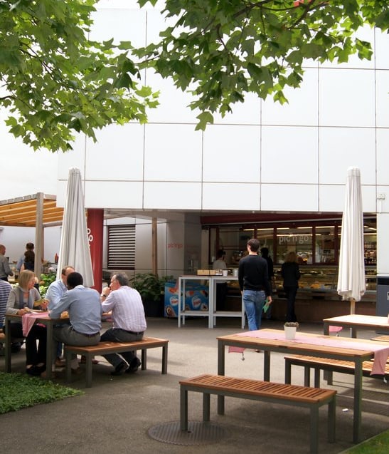 Menschen sitzen an Tischen im Freien neben einem Gebäude mit Bäumen.