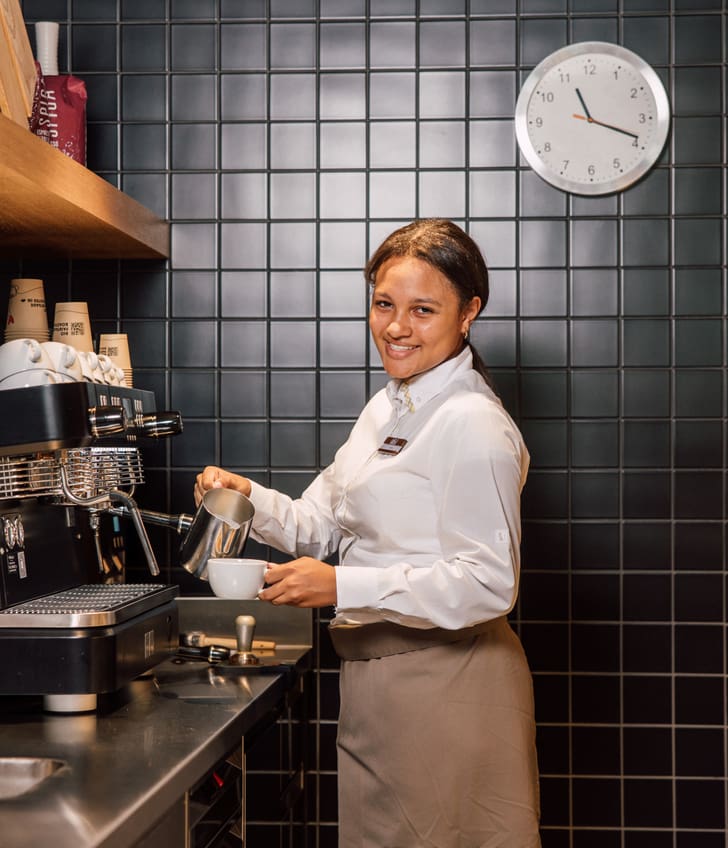 Barista macht Kaffee mit Espressomaschine, lächelnd, Wanduhr im Hintergrund.