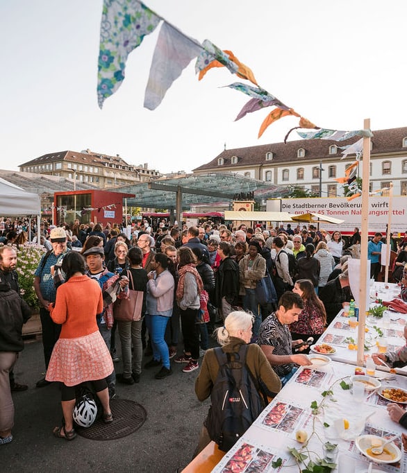 Versammlung von Menschen an einem Fest, im Vordergrund ein langer Tisch, an dem gegessen wird.