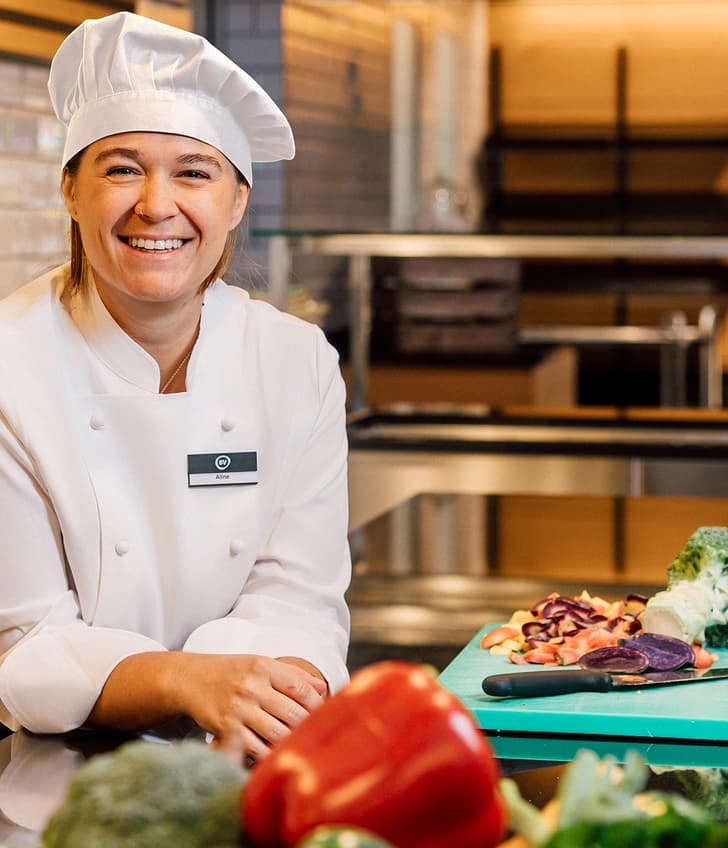 Köchin steht lächelnd in der Küche mit Gemüse auf Schneidebrett.