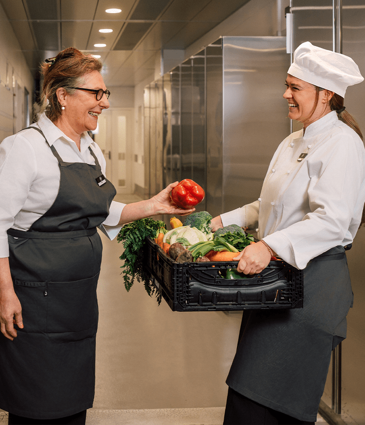 Zwei Köchinnen mit Gemüse in einer Küche lächelnd.