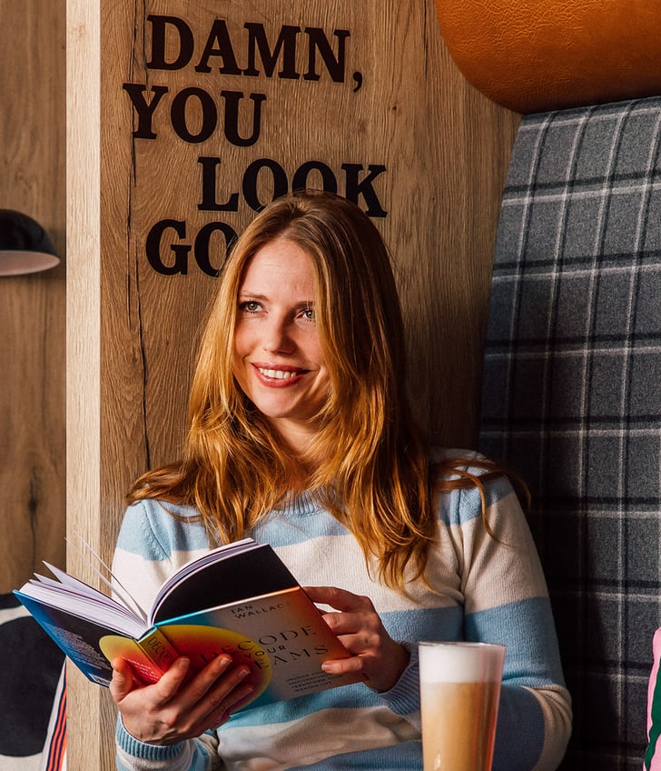 Frau mit Buch und Kaffee lächelnd in einer gemütlichen Ecke sitzend.
