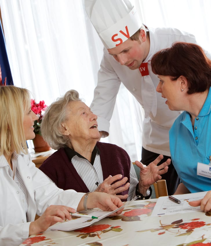 Ältere Frau mit Pflegepersonal und Koch in einem Gespräch am Tisch.