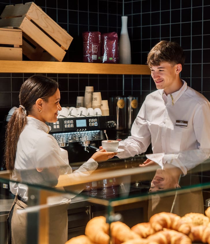 Zwei Mitarbeiter in einer Bäckerei mit Gebäck im Vordergrund, eine Frau überreicht eine Tasse an einen Mann.