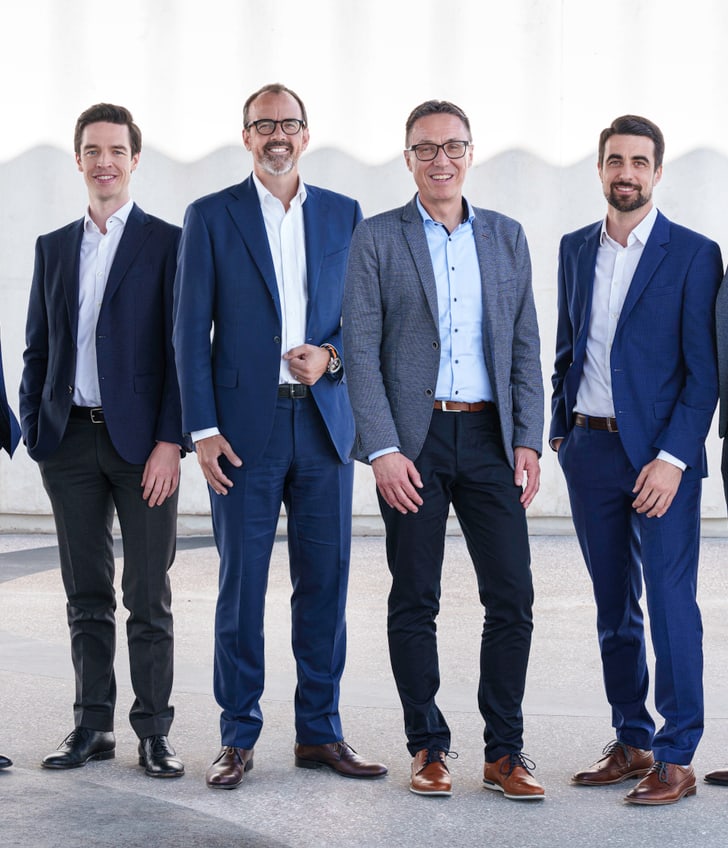 Sieben Männer in Anzügen stehen nebeneinander vor hellem Hintergrund.