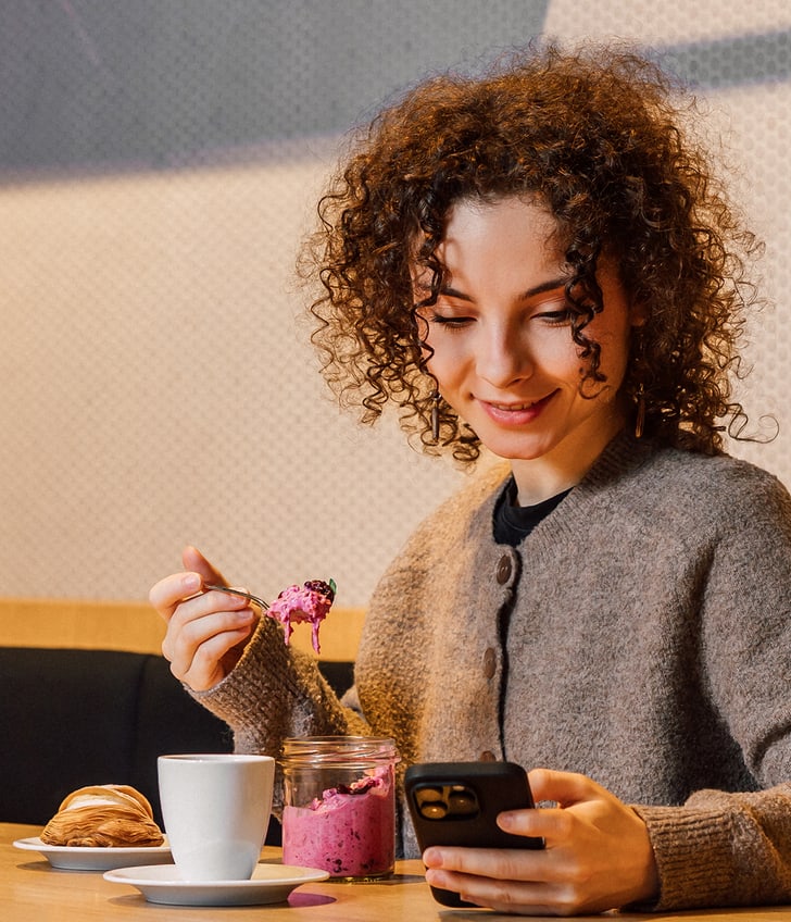 Frau isst Joghurt und schaut auf ihr Handy in einem Café.