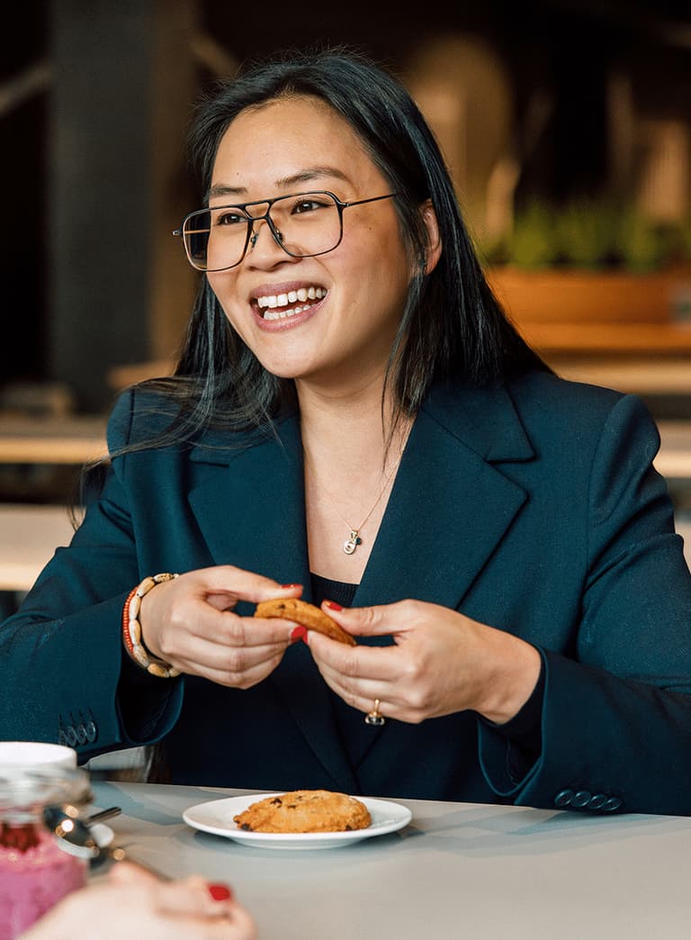 Frau in Blazer lächelnd und Keks haltend am Tisch sitzend.