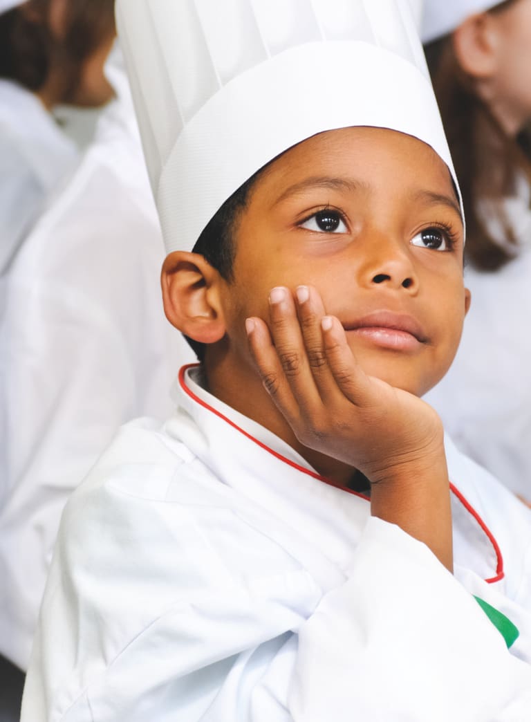 Kind mit einer weissen Kochuniform und Kochmütze sitzt in der Küche und denkt nach, andere Kinder sind im Hintergrund.