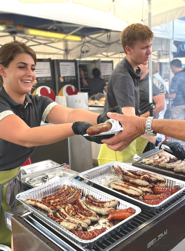 Mitarbeitede stehen an einem Grill bei einer Veranstaltung und geben frisch gebratene Würste an die Gäste heraus.