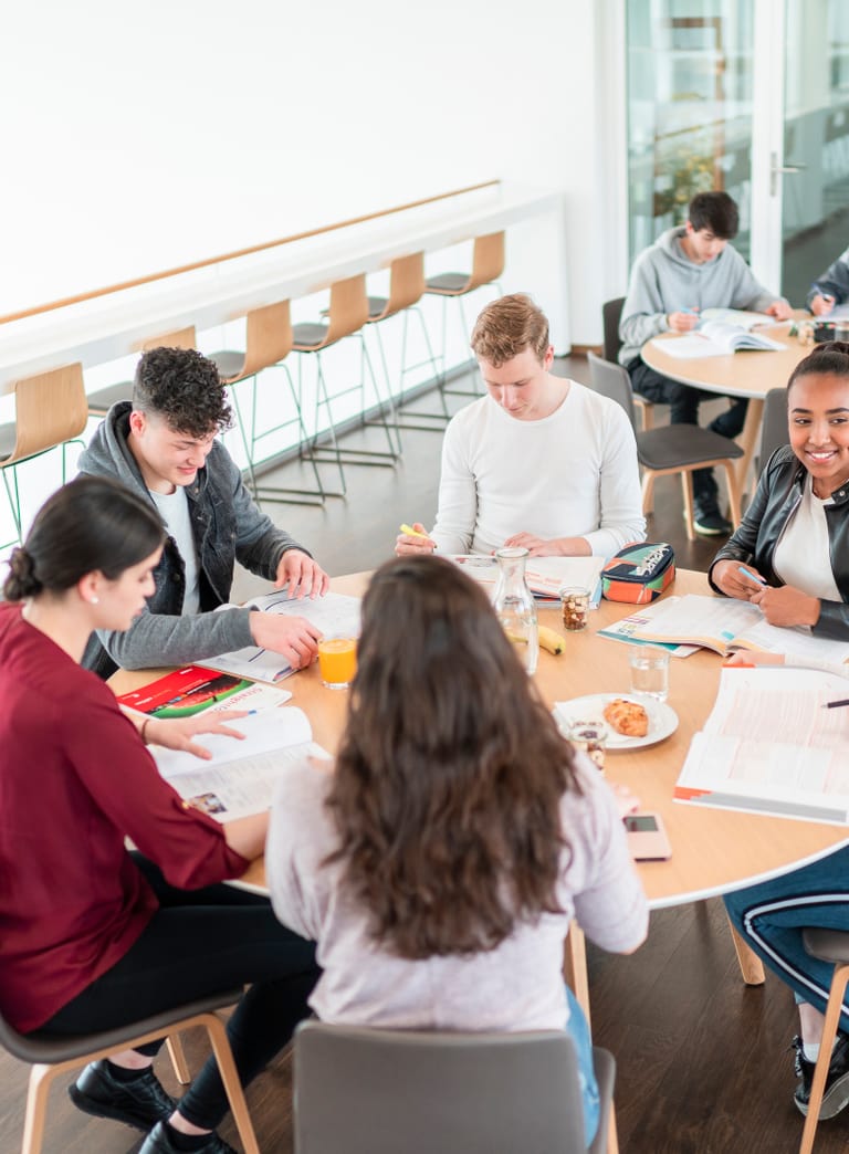 Schülerinnen und Schüler sitzen gemeinsam an einem runden Tisch in der Mensa ihrer Schule beim Lernen und geniessen dabei Snacks.