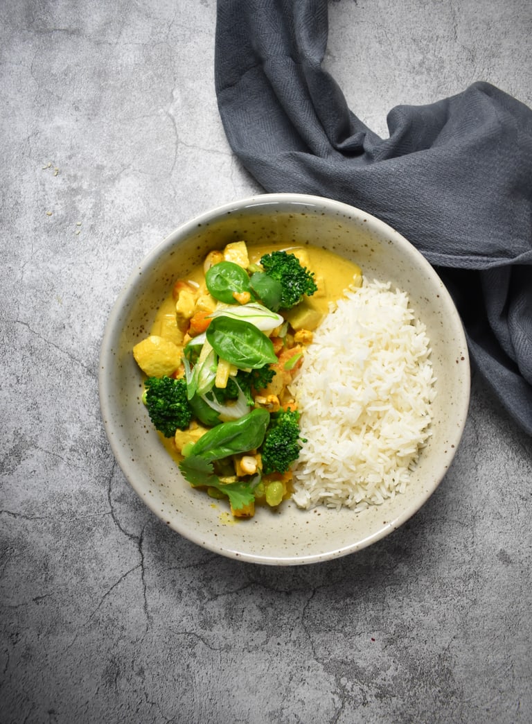 Teller mit Curry, Brokkoli und Reis auf grauem Untergrund.