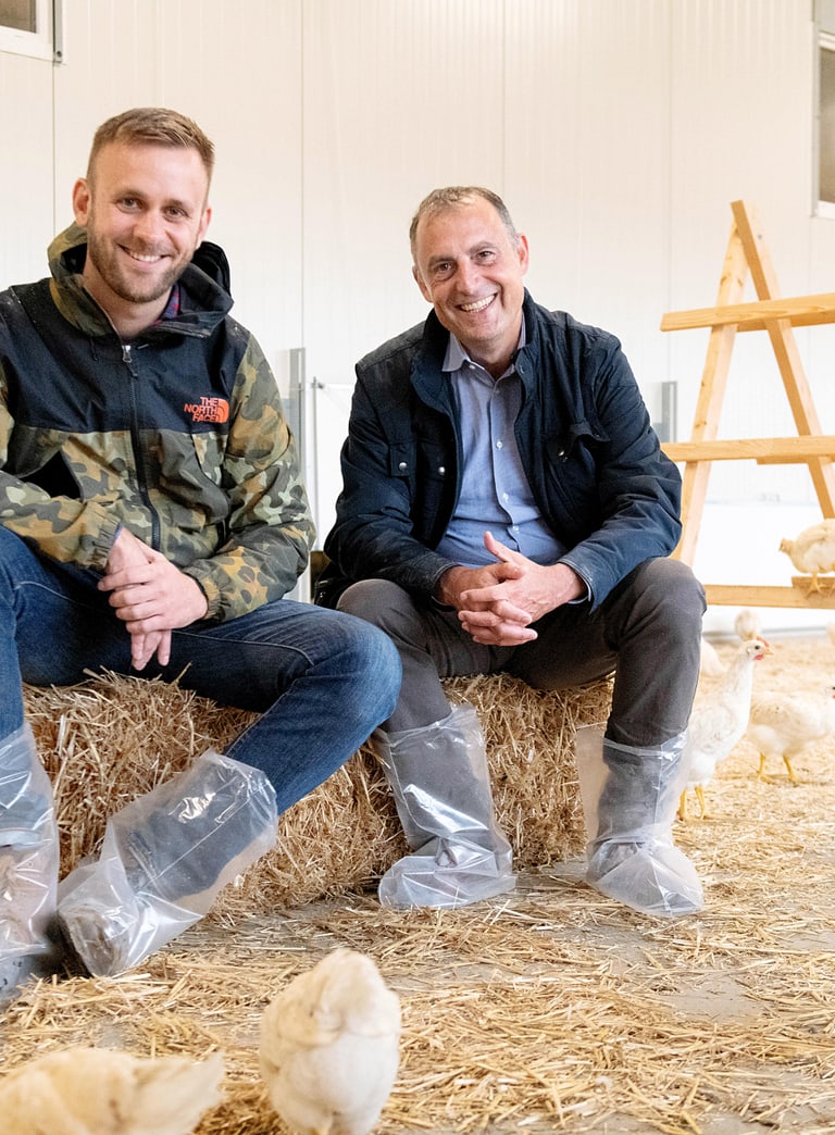 Foto von 2 sitzenden Männer auf einem Strohball in einem Hühnerstall. Im Bild sind auch noch ein paar Hühner