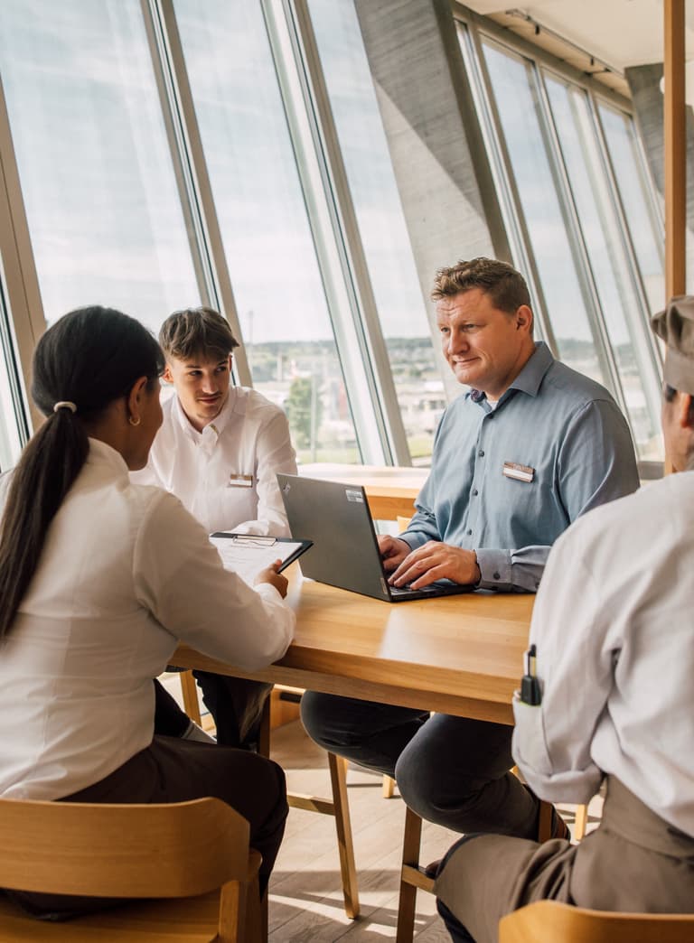 Leute in einem Meeting am Tisch mit Laptop und Fenstern im Hintergrund.