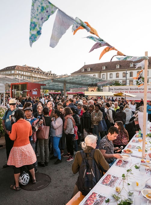 Versammlung von Menschen an einem Fest, im Vordergrund ein langer Tisch, an dem gegessen wird.