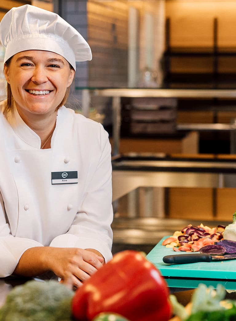 Köchin steht lächelnd in der Küche mit Gemüse auf Schneidebrett.