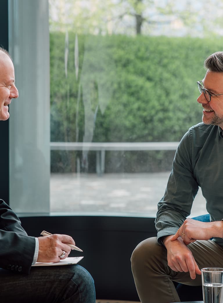 Zwei Männer unterhalten sich in modernem Büro mit grossem Fenster.