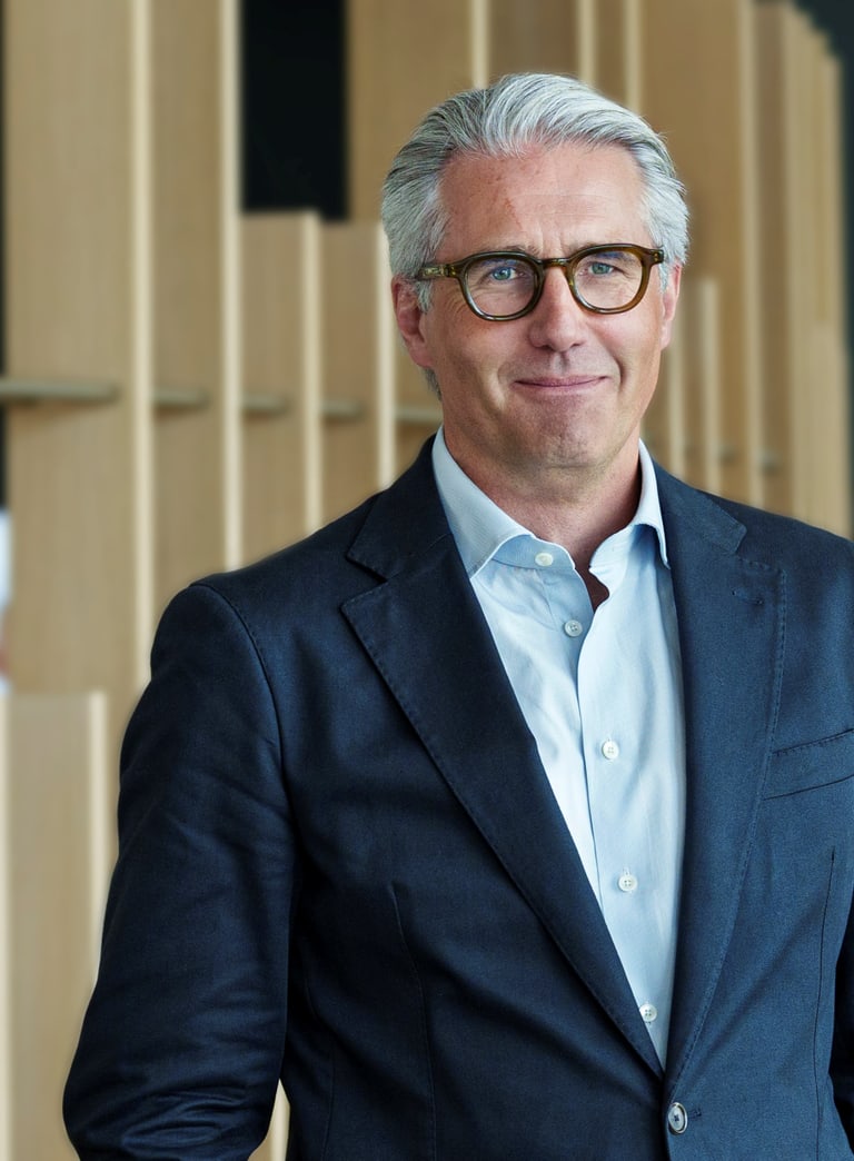 Mann mit grauem Haar und Brille in einem Anzug vor einem modernen Hintergrund.