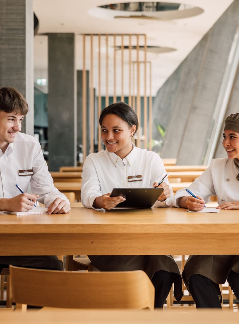 Drei Restaurantmitarbeiter in Uniform am Tisch sitzend, lächelnd.
