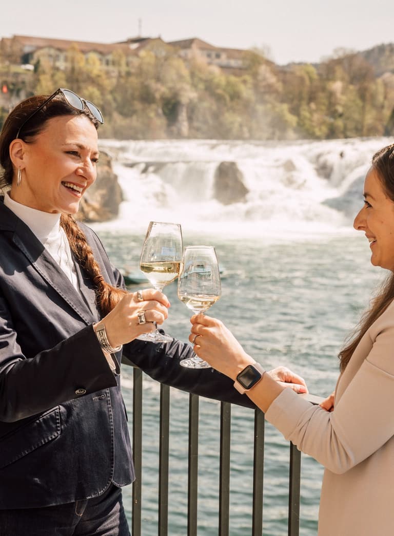 Zwei Frauen stossen mit Wein an vor Wasserfallkulisse.