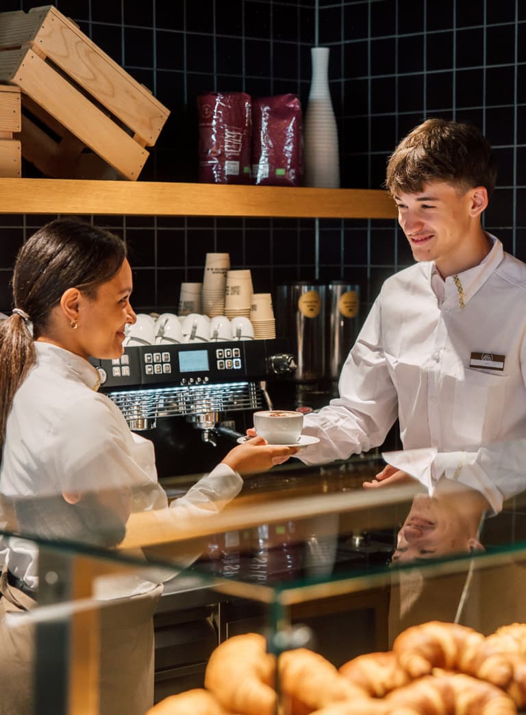 Zwei Mitarbeiter in einer Bäckerei mit Gebäck im Vordergrund, eine Frau überreicht eine Tasse an einen Mann.