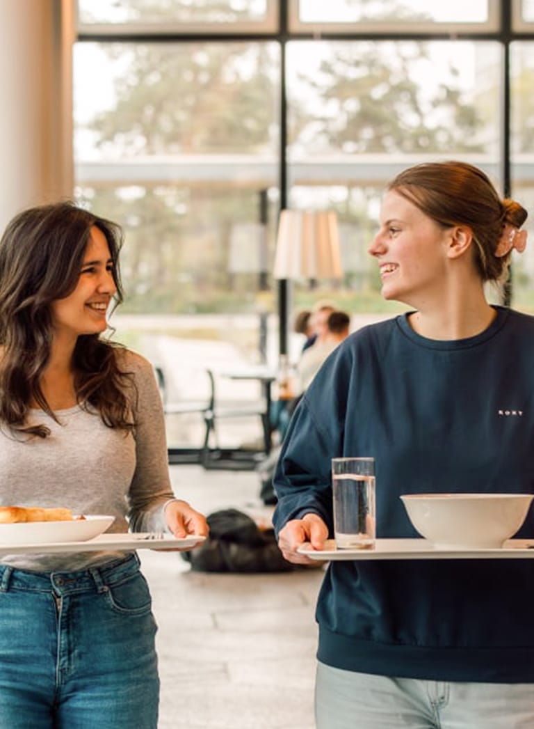 Zwei Frauen mit Tabletts im Essbereich, lächelnd.