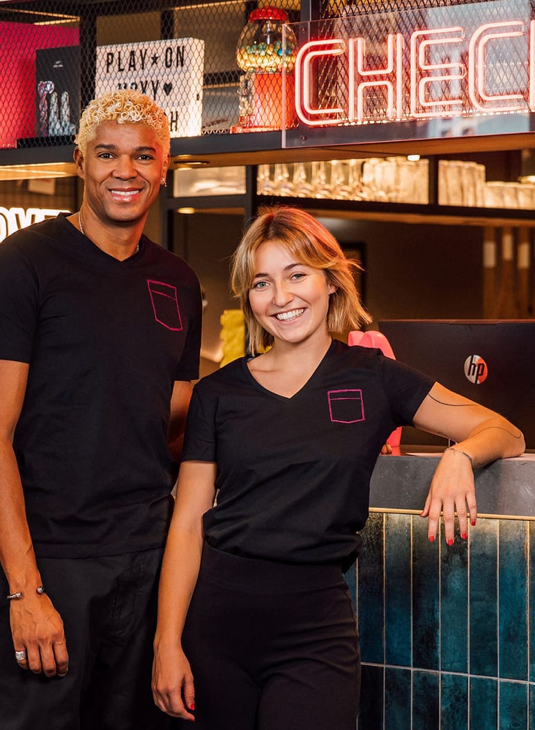 Zwei Personen in schwarzer Arbeitskleidung lächeln an einer Rezeption mit neonfarbener Dekoration.