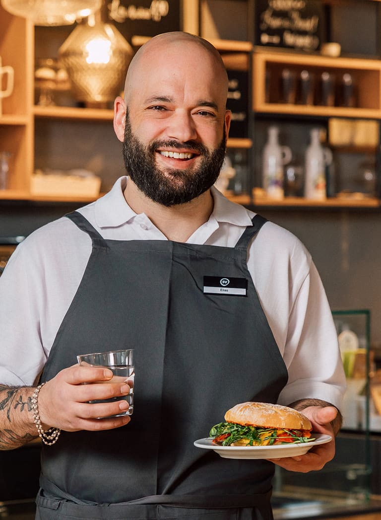 Lächelnder Mann in Schürze mit Sandwich und Getränk in Café.