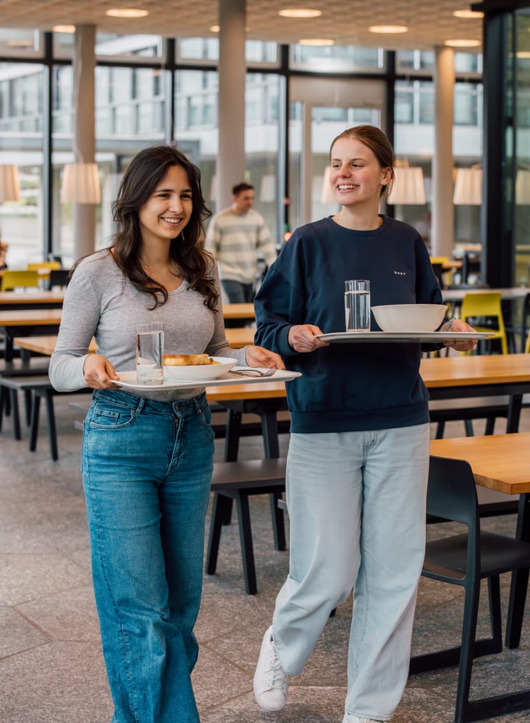 Zwei Personen mit Tabletts in einer Kantine gehend.