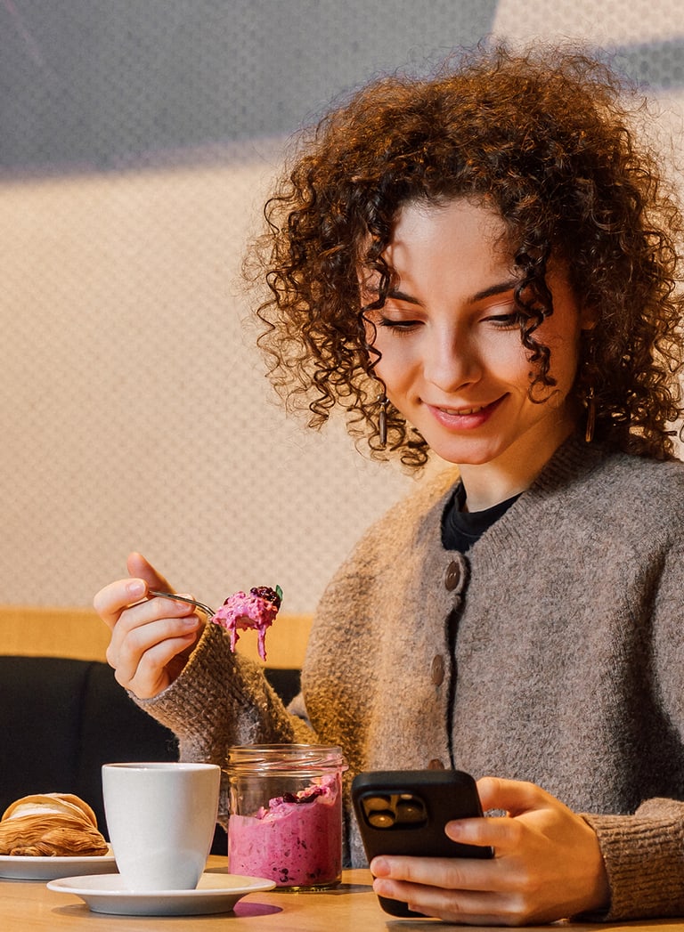 Frau isst Joghurt und schaut auf ihr Handy in einem Café.