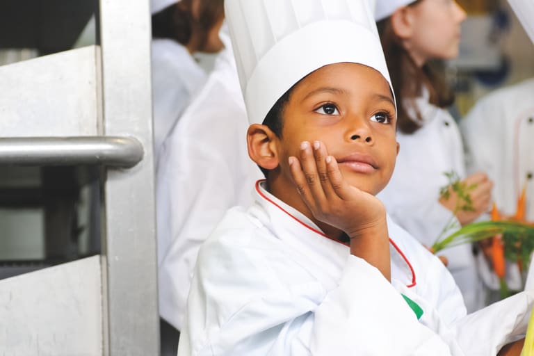Kind mit einer weissen Kochuniform und Kochmütze sitzt in der Küche und denkt nach, andere Kinder sind im Hintergrund.