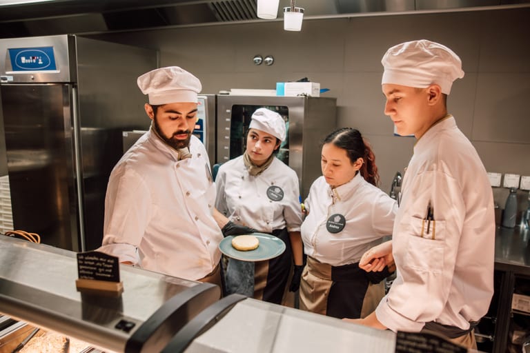Köche in einer Restaurantküche diskutieren über Präsentation eines Gerichts auf einem Teller.