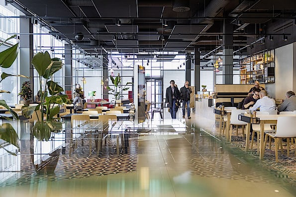 In einem modernen Mitarbeiterrestaurant mit Pflanzen und stylischem Mobiliar sitzen Gäste beim Mittagessen und laufen den Flur entlang.
