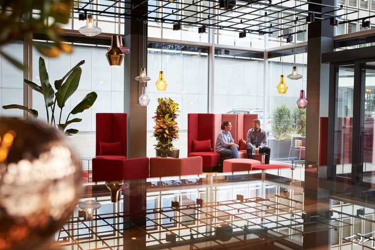 In einer modernen Lounge im Eingangsbereich eines Unternehmens unterhalten sich zwei Mitarbeitende.