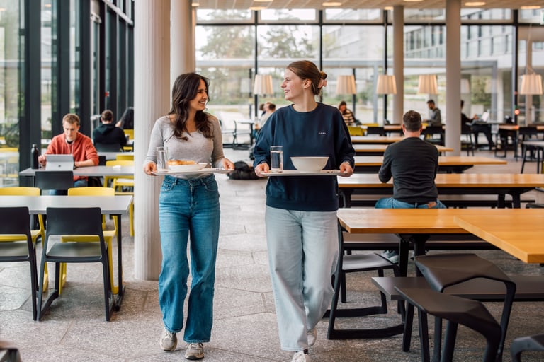 Zwei Frauen tragen Tabletts in einer Cafeteria.