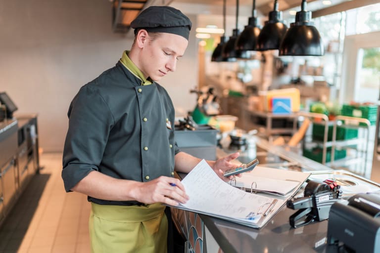 Ein Mitarbeiter in Kochuniform steht mit einer Checkliste an einem Counter.